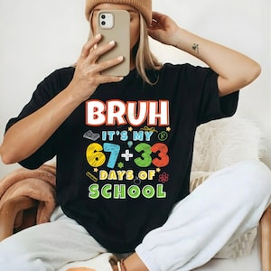 Op de afbeelding: Zwart T-shirt met de tekst "BRUH IT'S MY 67+33 DAYS OF SCHOOL" in kleurrijke letters. Het shirt heeft een ronde hals en korte mouwen. Het ontwerp bevat een boek, een ster en een wetenschappelijk symbool.