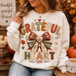 May include: A white long-sleeve sweatshirt featuring the text "GRATEFUL HEART" in a patchwork-style font. The design includes a large central bow with floral patterns, green gingham accents, and a small gold bell. Autumnal motifs like leaves and hearts surround the text.