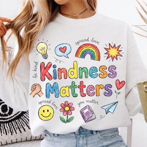 May include: White sweatshirt with a colorful graphic that reads "Kindness Matters." The design includes a rainbow, hearts, a smiley face, a flower, a lightbulb, and a paper airplane. The words "be kind," "spread love," and "you matter" are also included.