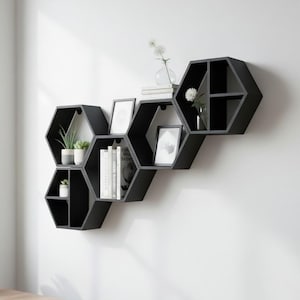 May include: A set of interconnected black hexagonal wall shelves arranged in a cluster. The shelves feature multiple compartments holding various items including small potted plants, framed pictures, and several books standing upright.