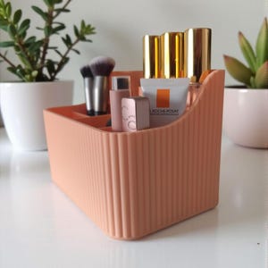 May include: A peach-colored, ribbed plastic organizer filled with makeup brushes, lipsticks, and skincare products. The organizer sits on a white surface, with potted plants in the background. The organizer has multiple compartments.