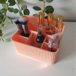 May include: A peach-colored, ribbed plastic organizer filled with makeup brushes, spray bottles, lipsticks, and tubes. The organizer has multiple compartments and is sitting on a white surface. Green leafy branches are in the background.