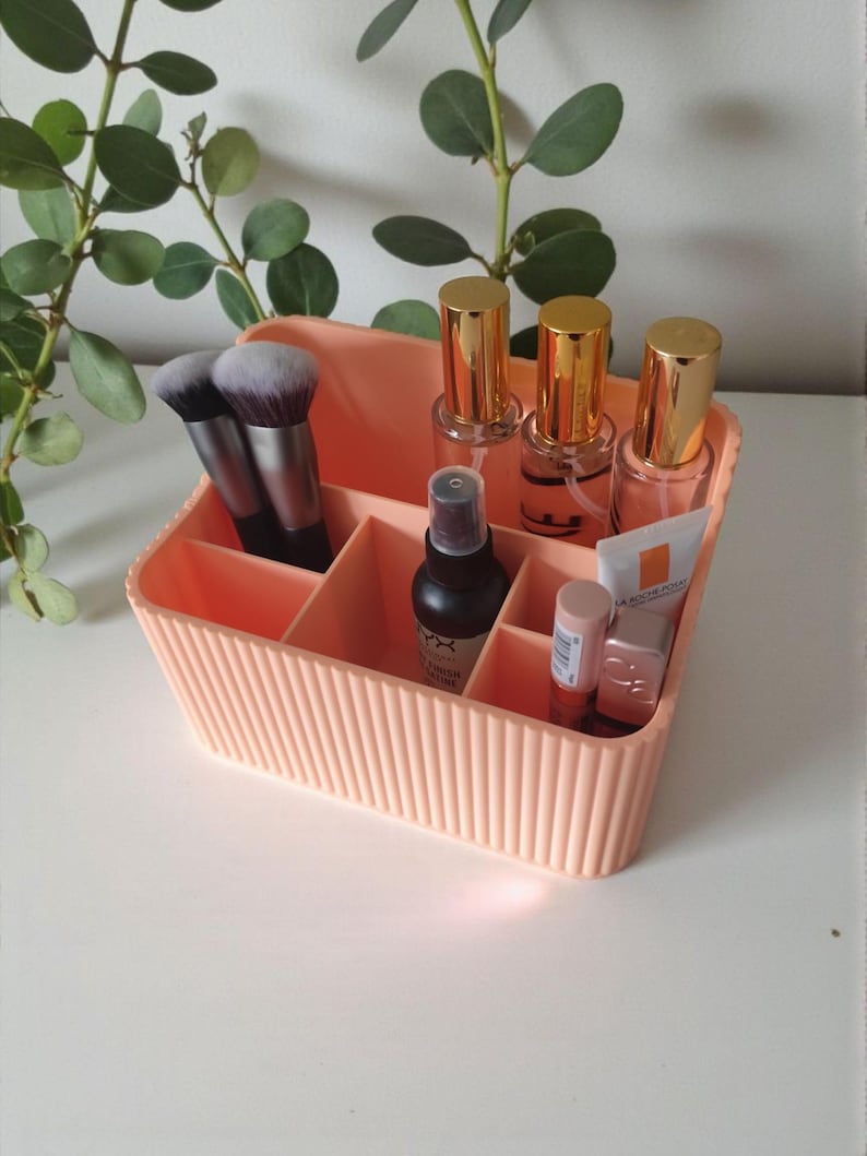 May include: A peach-colored, ribbed plastic organizer filled with makeup brushes, spray bottles, lipsticks, and tubes. The organizer has multiple compartments and is sitting on a white surface. Green leafy branches are in the background.