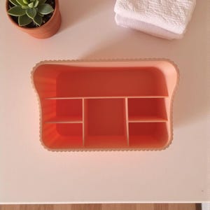May include: An orange plastic organizer with multiple compartments sits on a white surface. A small potted succulent and a folded white towel are also visible. The organizer has a scalloped edge.
