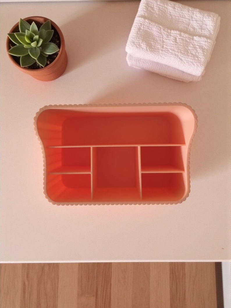 May include: An orange plastic organizer with multiple compartments sits on a white surface. A small potted succulent and a folded white towel are also visible. The organizer has a scalloped edge.