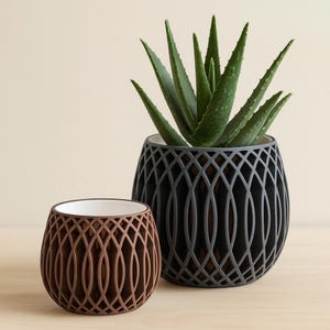 May include: Two rounded planters with a lattice-style outer shell and a smooth white inner liner. The larger dark gray planter contains a green aloe vera plant, while the smaller brown planter is empty. Both feature a repeating geometric pattern.