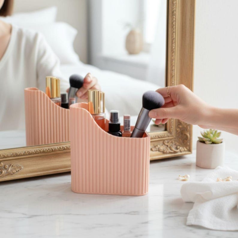 May include: A peach-colored, ribbed cosmetic organizer holding makeup brushes and bottles. The organizer sits on a marble surface, with a mirror reflecting the scene. A hand reaches for a makeup brush. A small succulent plant is visible.