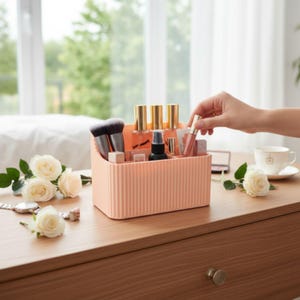 May include: A peach-colored, ribbed makeup organizer filled with beauty products, including brushes, lipsticks, and perfume bottles. A hand reaches for a lip gloss. White roses and a watch are on the wooden surface.