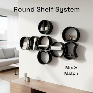 May include: Black round wall shelves arranged in a unique pattern, holding decorative items. The shelves are mounted on a white wall. The text "Round Shelf System" and "Mix & Match" are displayed.