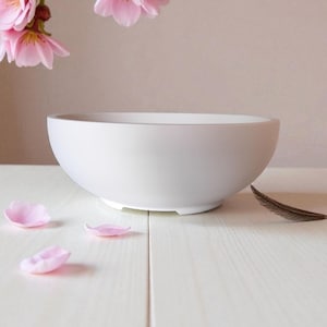 May include: A white ceramic bowl with a rounded shape, set on a white surface. Pink flower petals and a brown feather are scattered around the bowl. Pink flowers hang in the background, creating a serene, minimalist aesthetic.