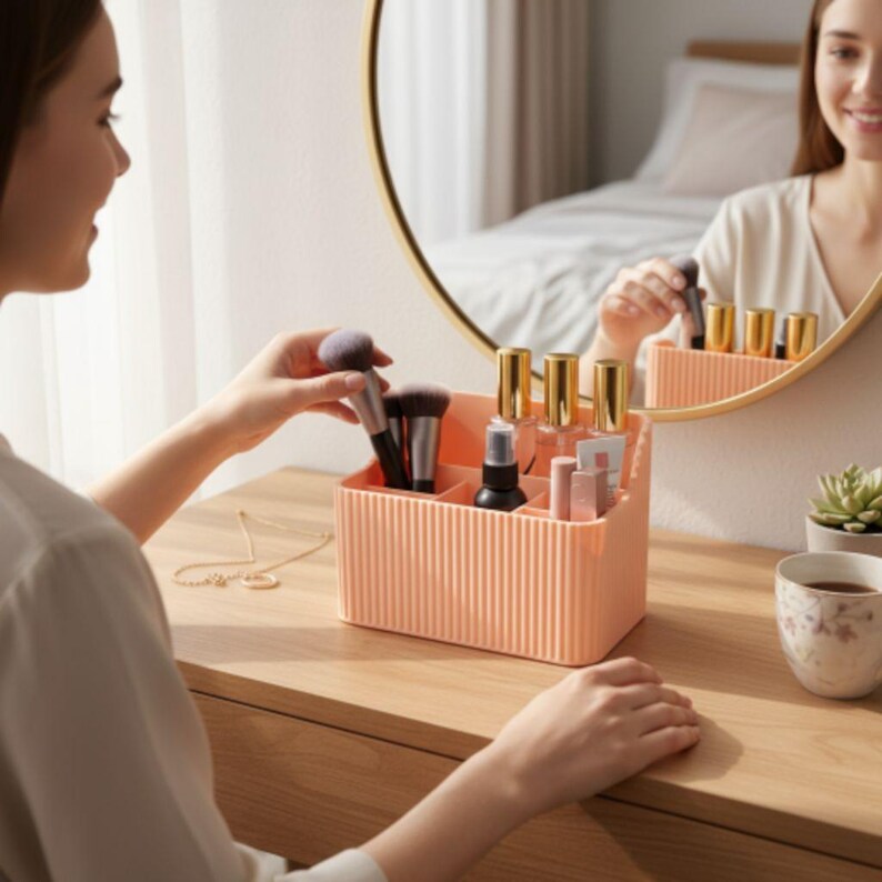 May include: A peach-colored, ribbed makeup organizer with multiple compartments. The organizer holds makeup brushes, bottles, and tubes. A woman is seen reaching for a brush. A gold-framed mirror reflects the organizer and other items.