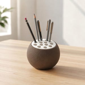 May include: A spherical desk organizer with a textured brown exterior and a flat white top featuring multiple circular holes. Several writing instruments, including pencils and pens in shades of black, grey, and silver, are inserted into the slots.
