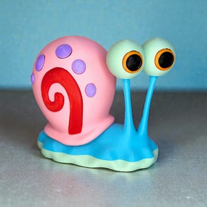 May include: A snail figurine featuring a bright pink shell with a red spiral pattern and purple circular spots. The snail has a light blue body, a pale green base, and two tall stalks topped with pale green eyes, orange irises, and black pupils.