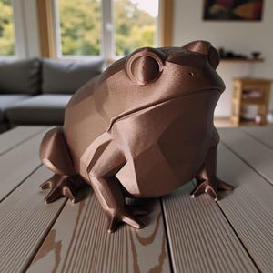 May include: A bronze-colored, geometric frog sculpture. The frog has a stylized, faceted design with visible ridges and a smooth finish. It is posed on a wooden surface, showcasing its detailed legs and body. The sculpture is a decorative item, suitable for home decor.