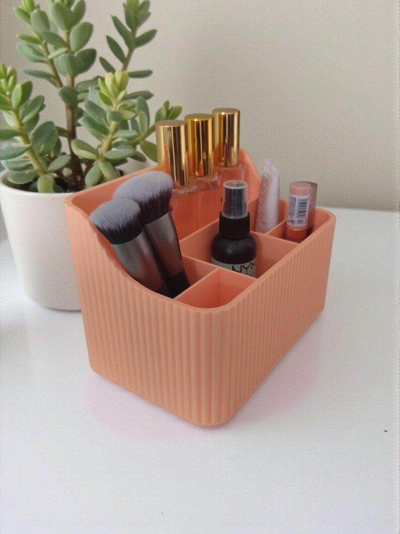 May include: A peach-colored, ribbed makeup organizer with multiple compartments. It holds makeup brushes, bottles with gold caps, and other beauty products. A small succulent plant in a white pot is in the background.