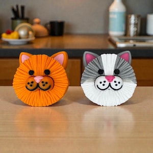 May include: Two cat-shaped decorative items. One is orange with pink accents, the other is gray, white, and pink. Both have black eyes and a pink nose. The items are on a light brown surface.