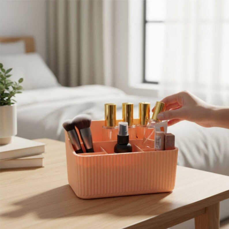 May include: A peach-colored, ribbed cosmetic organizer with multiple compartments. The organizer holds makeup brushes, perfume bottles, and other beauty products. A hand reaches for a bottle with a gold cap. The organizer sits on a wooden table.
