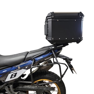 Peut inclure: Coffre supérieur de moto noir monté sur une moto bleue et noire. Le coffre supérieur est rectangulaire avec des coins arrondis et une garniture argentée. La moto a une selle noire et une roue dorée.