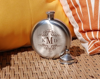May include: A silver-coloured, round flask with a screw-on cap and a separate funnel. The flask features an engraved monogram design with the letters "KMD". The flask is sitting on a woven surface, with a yellow cushion and an orange and white striped towel in the background.