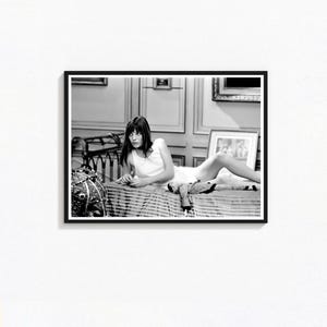 May include: Framed black and white print showing a woman in a white dress lying on a striped bed. The photograph has a vintage feel. The background features framed pictures and decorative details, creating a classic aesthetic.