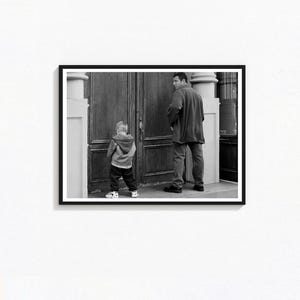 Puede incluir: Fotografía enmarcada en blanco y negro de un hombre y un niño cerca de una puerta de madera. El hombre lleva chaqueta y vaqueros, el niño una chaqueta con capucha y pantalones. La foto está tomada al aire libre.