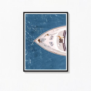 May include: An aerial view of a white yacht with people on board, framed in a black border. The yacht is on a blue sea. The boat has a pointed bow and a deck with seating and a small table. The image is a print.