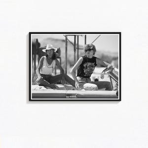 Puede incluir: Fotografía enmarcada en blanco y negro que muestra a dos personas sentadas en un coche. Una persona lleva un sombrero de vaquero y una camiseta sin mangas, la otra una camiseta gráfica y gafas de sol. El fondo muestra un paisaje desértico.