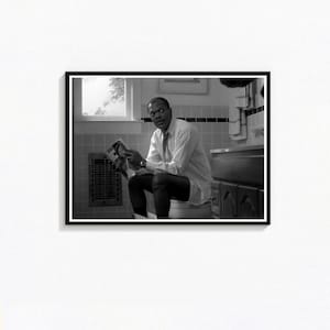 May include: Black and white framed photograph of a person sitting on a toilet, reading a magazine. The person is wearing a white dress shirt and tie. The bathroom has a window, a radiator, and a sink.