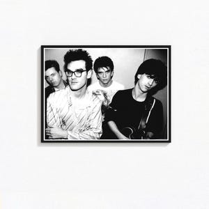May include: Black and white framed photograph of a band. Four men are in the image. One man is wearing glasses and a striped shirt. Another man is wearing a shirt with the text "The Smiths". The other two men are wearing black shirts.
