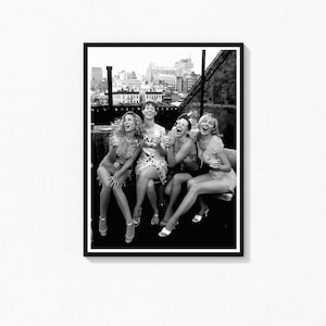 May include: Black and white photograph of five women laughing and sitting together on a rooftop. They are dressed in various outfits, including dresses and shorts. The background shows a cityscape.