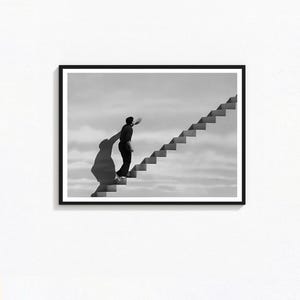 May include: Monochrome framed print depicting a person climbing a staircase against a cloudy sky. The image has a surreal feel, with a large shadow extending from the person. The artwork is a study in perspective and form.
