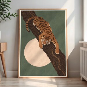 Op de afbeelding: Een ingelijste kunstdruk met een oranje en zwart gestreepte tijger die rust op een donkerbruine boomtak. De achtergrond is een effen gedempte groene kleur met een grote, lichtbeige cirkelvorm achter de boomtak geplaatst.