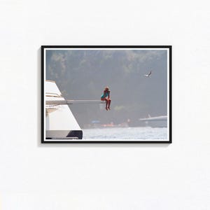 May include: Framed print showing a person in a blue swimsuit seated on the edge of a yacht. The background includes water, mountains, and a bird in flight. The yacht is white, set against a hazy backdrop.