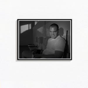 May include: Monochrome framed print depicting a man in a white t-shirt seated on a toilet, with a rifle against the wall. The man's face shows an intense expression. The background shows a row of toilets and a window.