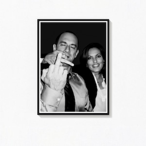 May include: Black and white framed photograph of two people. One person is giving the middle finger, while the other is smiling. The photograph is mounted on a white wall.