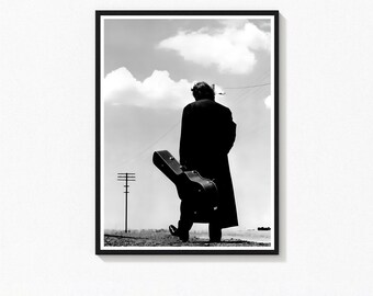 May include: Black and white framed print of a person walking away from the viewer, carrying a guitar case. The person is wearing a long coat and walking on a gravel path. A telephone pole and cloudy sky are in the background.