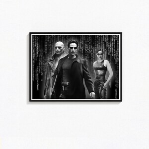 Peut inclure: Affiche encadrée en noir et blanc représentant trois personnages du film "Matrix". La figure centrale porte un long manteau noir et des lunettes de soleil. Le fond présente un effet de pluie numérique.