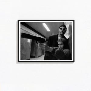 Puede incluir: Impresión enmarcada en blanco y negro que presenta a un hombre con gafas de sol sosteniendo una pistola, con una persona joven detrás de él. La imagen es un fotograma de una película, centrado en los personajes y sus expresiones.