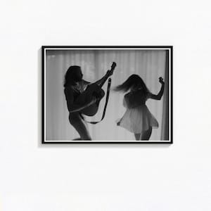 以下が含まれることがあります： ギターを弾く男性と踊る人物の白黒フレームプリント。男性はストラップ付きのギターを持っており、人物はドレスを着ています。背景はシアーなカーテンです。