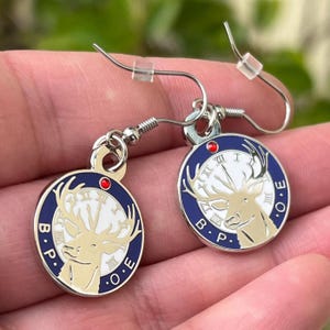 May include: A pair of silver-tone earrings with a circular pendant featuring a white clock face, a gold-colored deer, and blue and white accents. The letters "B.P.O.E." are around the edge. Each earring has a red accent.
