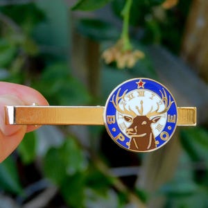 Pode incluir: Grampo de gravata dourado com um emblema circular com uma cabeça de veado, chifres e as letras "B.P.O.E." em um design azul e branco. O emblema tem uma estrela vermelha no topo.