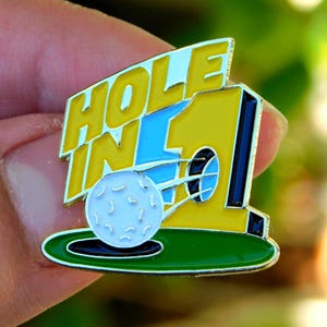 May include: A golf-themed pin with the words "HOLE IN 1" in yellow and white lettering. The pin features a golf ball on a green surface, with a yellow and blue background. The pin is held by a person.