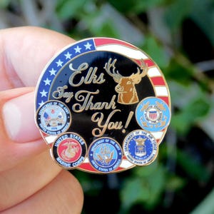 May include: A round, patriotic pin with the text "Elks Say Thank You!" and images of the U.S. military branches. The pin features a black center with a gold elk, surrounded by a red, white, and blue border.