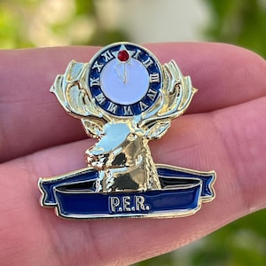Puede incluir: Broche metálico dorado con la cabeza de un ciervo con cuernos, una esfera de reloj y una cinta azul con las letras "P.E.R.". El reloj tiene números romanos y una gema roja. Accesorio decorativo.