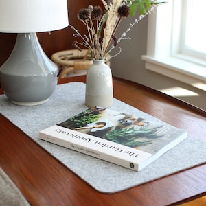K&ouml;nnte beinhalten: Ein grauer Filz-Tischl&auml;ufer mit einem Buch mit dem Titel "The Garden Apothecary" darauf. Eine graue Keramiklampe und eine kleine Vase mit getrockneten Blumen stehen auf dem Tisch.