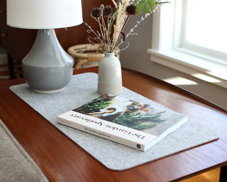 K&ouml;nnte beinhalten: Ein grauer Filz-Tischl&auml;ufer mit einem Buch mit dem Titel "The Garden Apothecary" darauf. Eine graue Keramiklampe und eine kleine Vase mit getrockneten Blumen stehen auf dem Tisch.