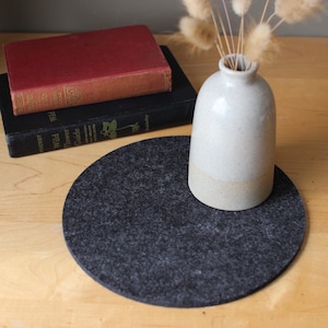May include: A gray felt coaster with a white ceramic vase with dried flowers on top. The vase is round and has a textured surface.
