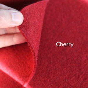 May include: A piece of cherry red felt fabric with a soft, dense, and slightly fuzzy texture. The material is folded over to reveal its thickness and consistent color. The word Cherry is printed in white text on the right side of the fabric.