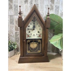 Puede incluir: Un reloj regulador de madera con una parte superior triangular y pináculos decorativos. La esfera del reloj es blanca con números y manecillas negras y detalles florales. El péndulo es visible a través de una puerta de cristal.