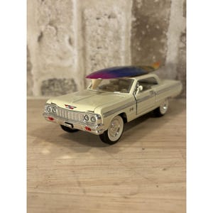 May include: A cream-colored classic car with a surfboard on top. The car has white-wall tires and chrome detailing. The surfboard is painted with a gradient of colors, including purple, blue, and orange. The car is a vintage model.